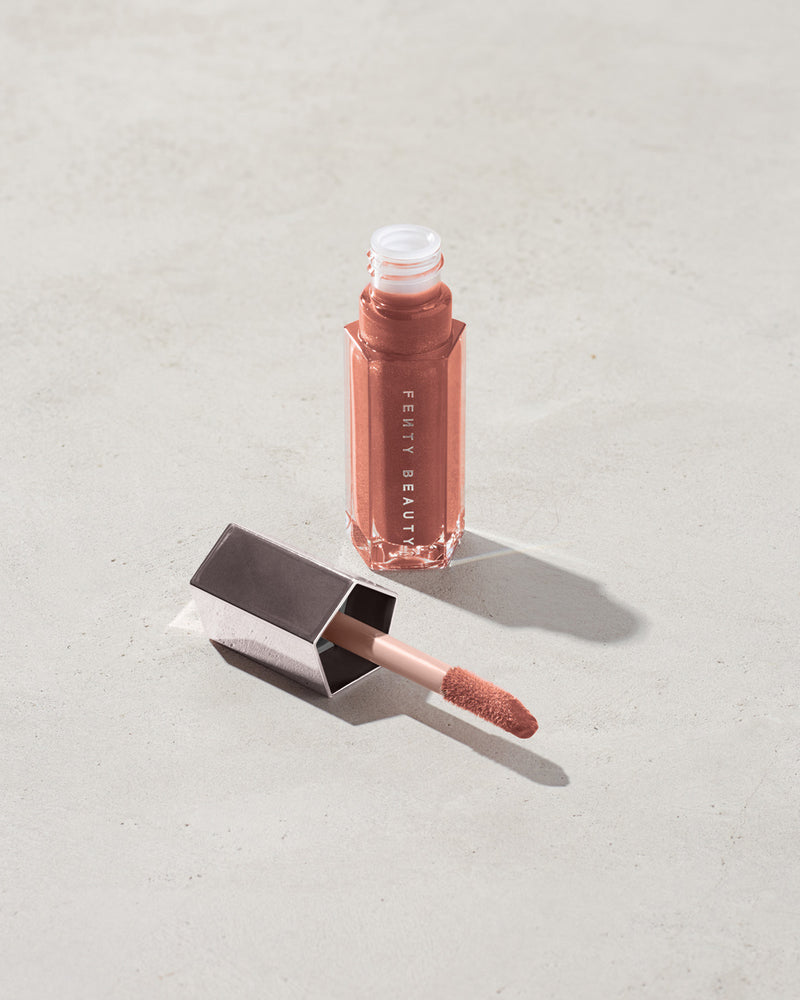 An open tube of Gloss Bomb Universal Lip Luminizer in the shade Fenty Glow on a grey background.